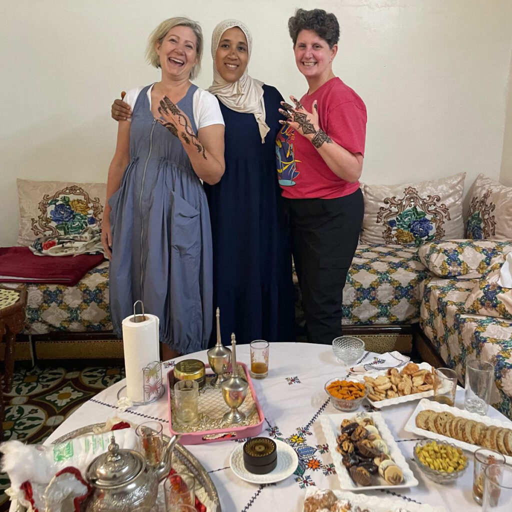 Three women stand smiling in a living room behind a table set with tea, snacks, and sweets. Two of them have henna designs on their hands. The setting appears warm and casual.