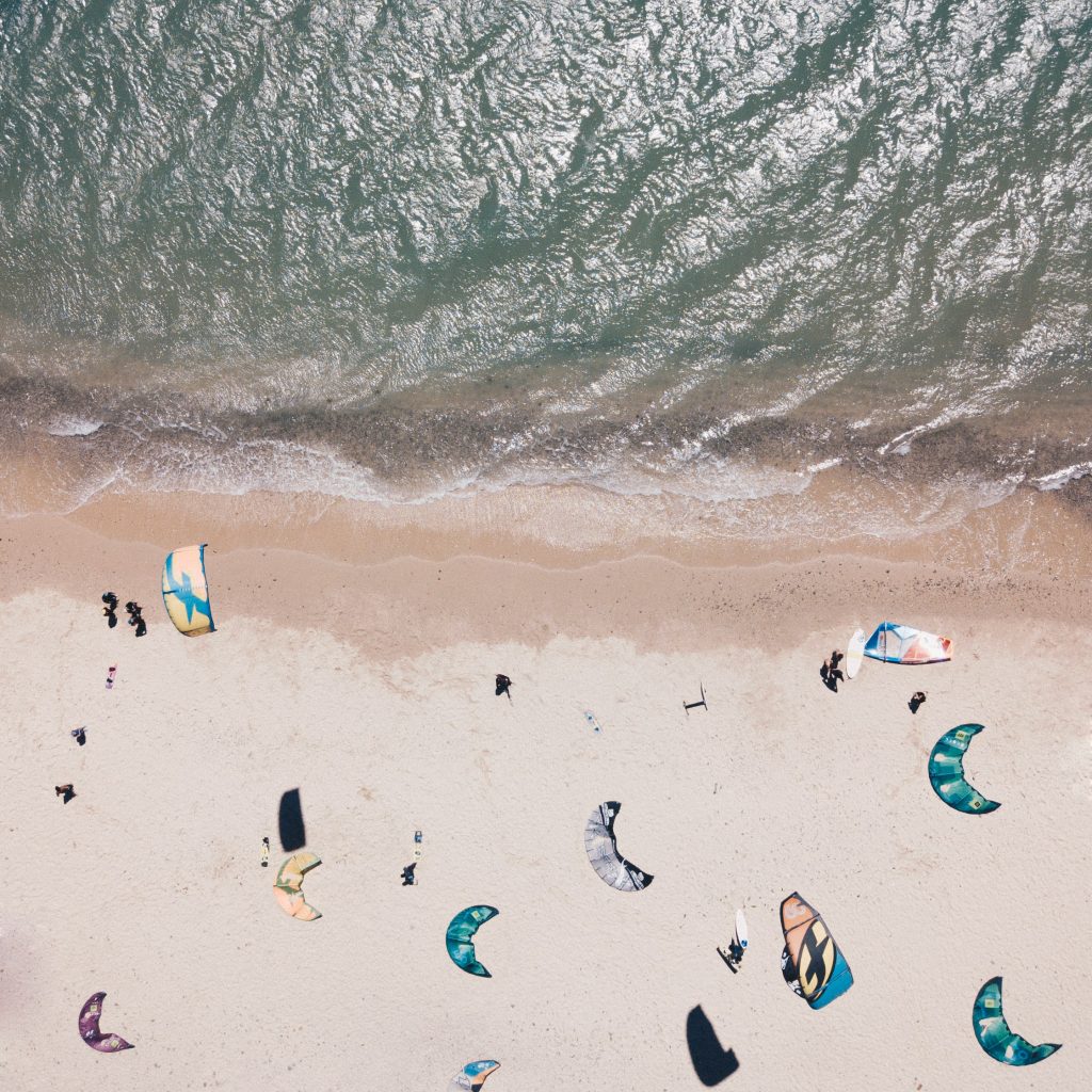 Aerial view of a sandy beach with people and colorful kitesurfing kites lying on the shore near the water’s edge.
