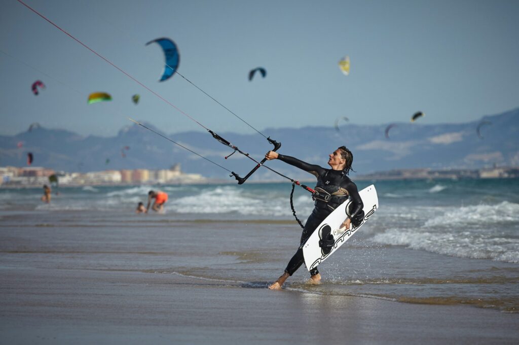 Person in a wetsuit walks on the beach carrying a kiteboard and holding kite control lines, with other kite surfers and mountains in the background.