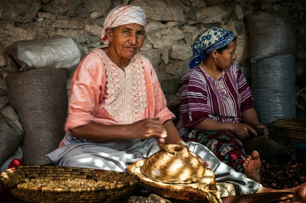 Two women sit indoors; one grinds argan nuts with a stone mill while the other works beside her. Baskets and sacks of nuts are around them. Light filters through the roof above.
