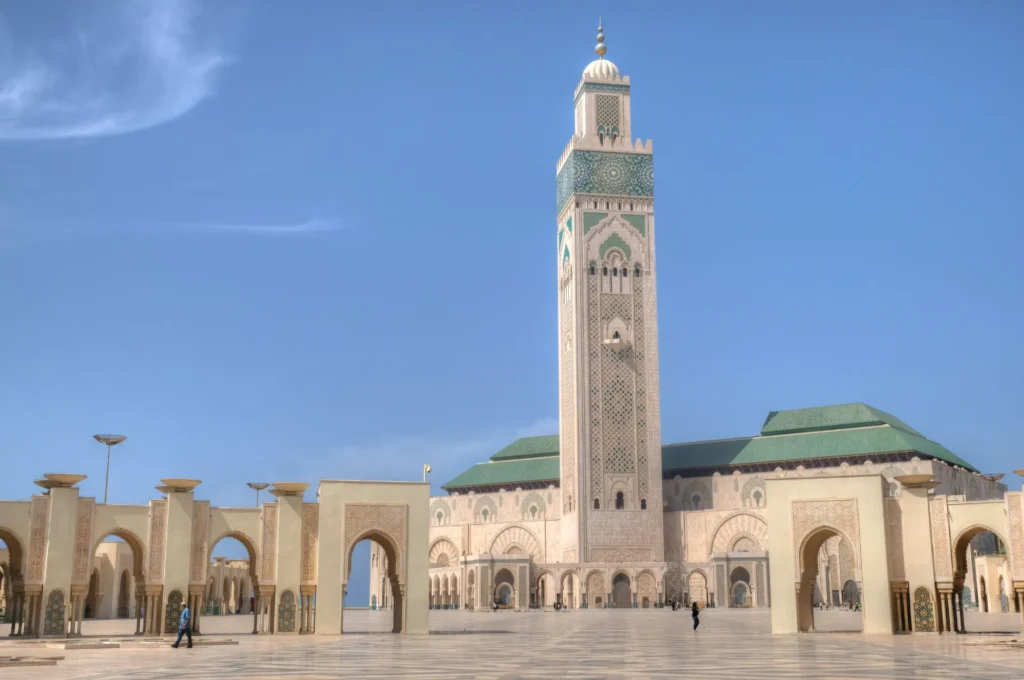 The image shows the Hassan II Mosque in Casablanca, Morocco, featuring its tall minaret, arched entrances, and a spacious courtyard under a clear blue sky.