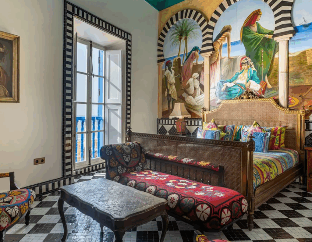 A colorful bedroom with a mural of figures and palm trees, patterned textiles on the bed and furniture, and black-and-white tiled floors and archways.