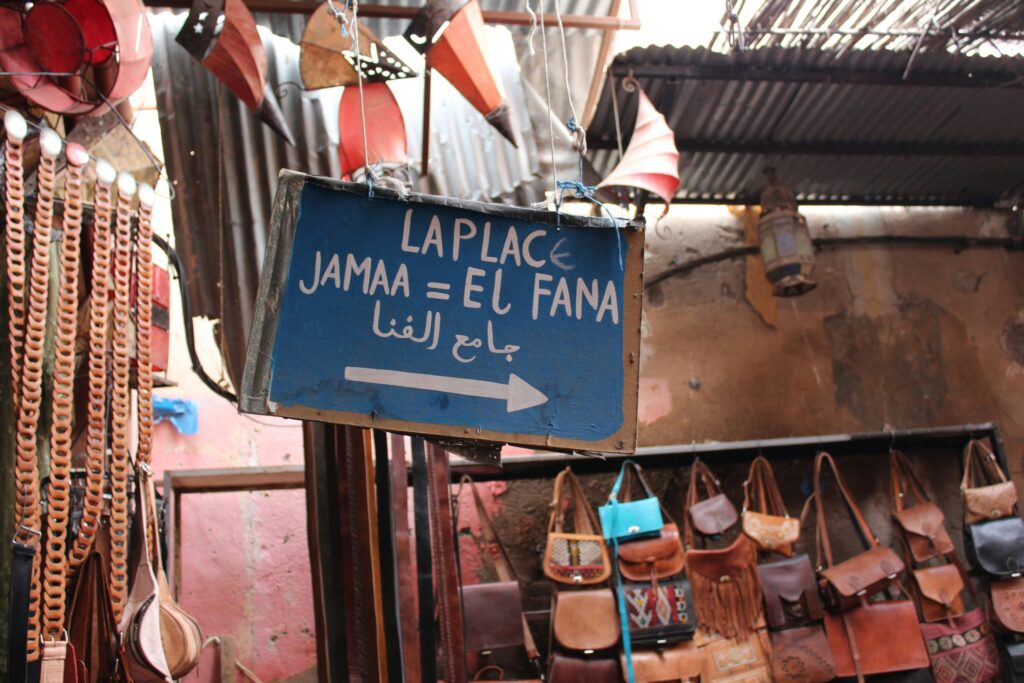 A blue sign with white text directs to La Place Jamaa El Fana in Arabic and French. Leather bags hang below the sign in a market setting.
