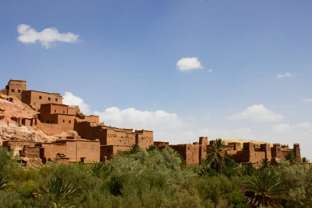 A historic fortified village with earth-toned buildings stands on a hillside, surrounded by greenery under a blue sky with scattered clouds.