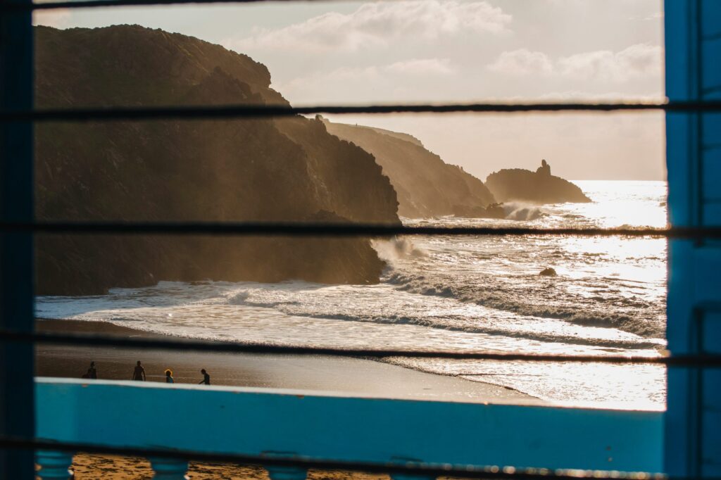 The photo shows a rocky coastline with waves crashing on the shore, viewed through horizontal bars, with several people walking on the sandy beach below.