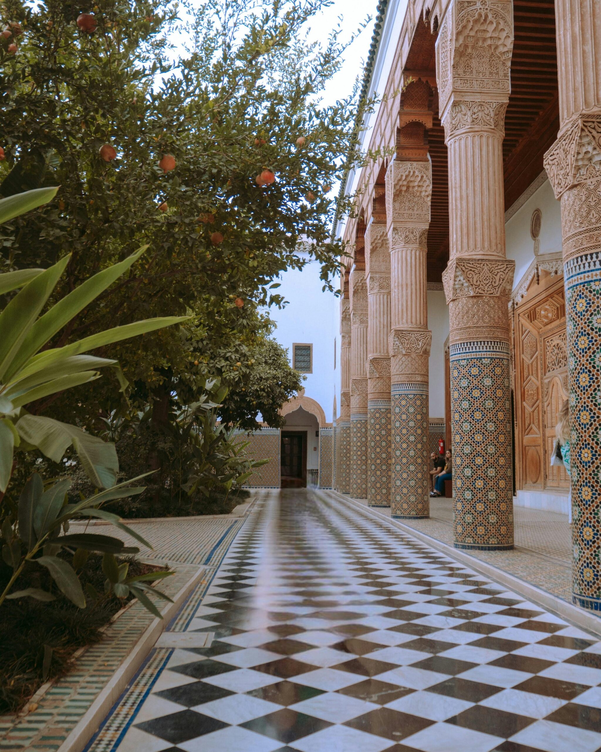 A tiled walkway with black and white checkerboard pattern runs alongside ornately carved columns and lush greenery in a traditional courtyard.