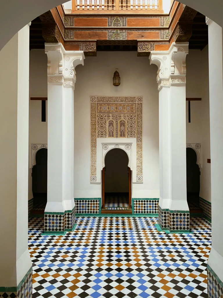 Traditional Moroccan interior with ornate columns, arched doorway, geometric tile floor, and intricate wood and plaster detailing on walls and ceiling.