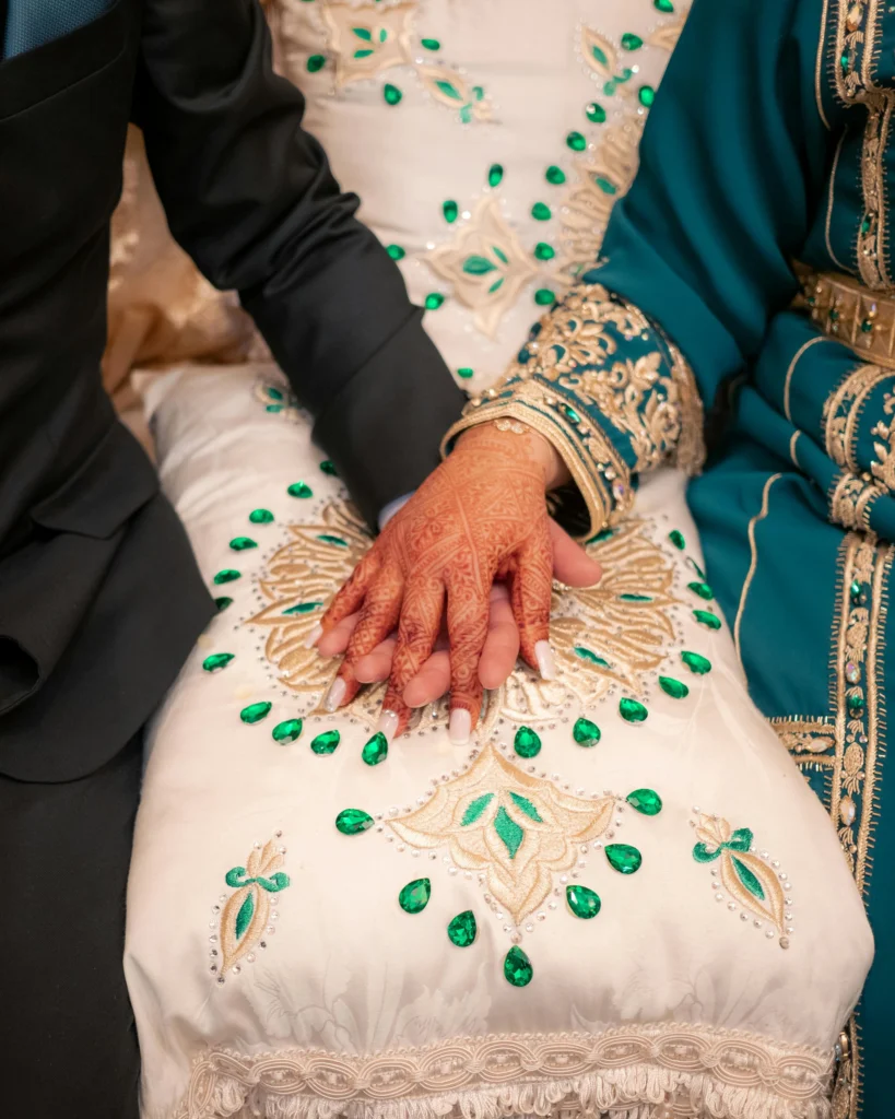 Two people sit close together, holding hands on a white cloth decorated with green and gold embroidery. One wears a dark suit, the other a green embroidered outfit with henna on their hand.