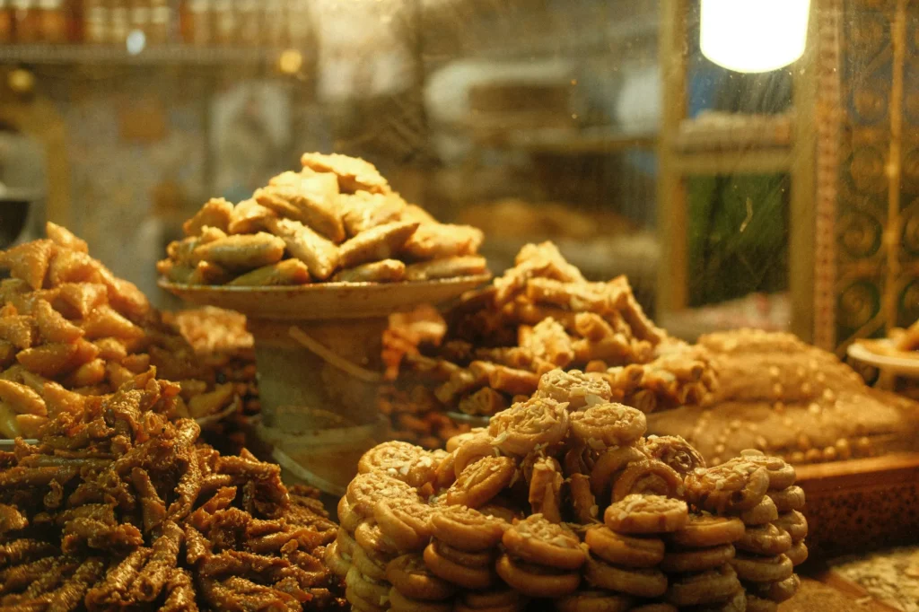 Various types of pastries and sweets are stacked on trays and dishes behind a glass counter in a bakery or market.
