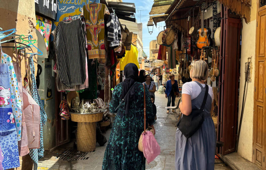 Two women walk through a narrow marketplace, lined with hanging clothes, musical instruments, and various goods, exploring the vibrant scene like Siham Lahmine, founder of Morocco Travel Organizer.