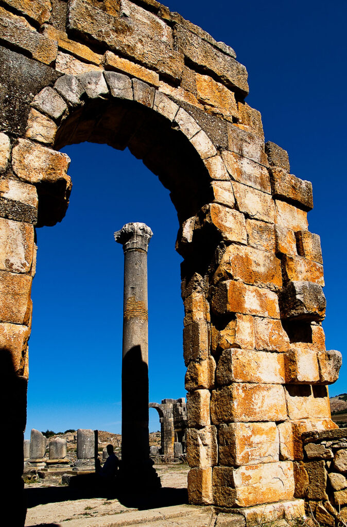 Ancient stone archway frames a single standing column amid other ruins under a clear blue sky.
