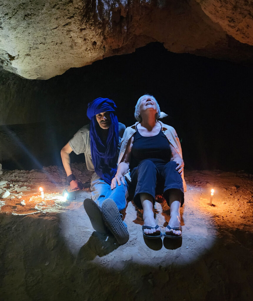 Two people sit on the ground inside a dimly-lit cave, illuminated by three small candles; one person wears a blue headscarf, the other smiles and looks up.