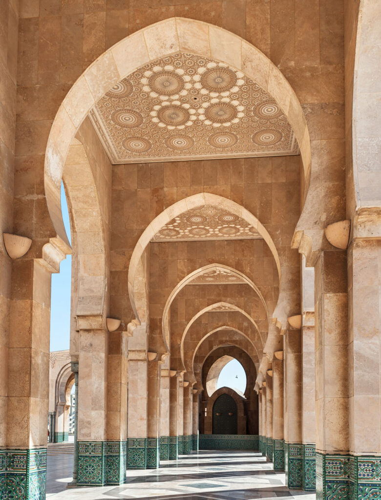 Stone arches and ornate ceilings with geometric patterns in a corridor of a mosque, featuring green tile details at the base of columns and sunlight streaming through.