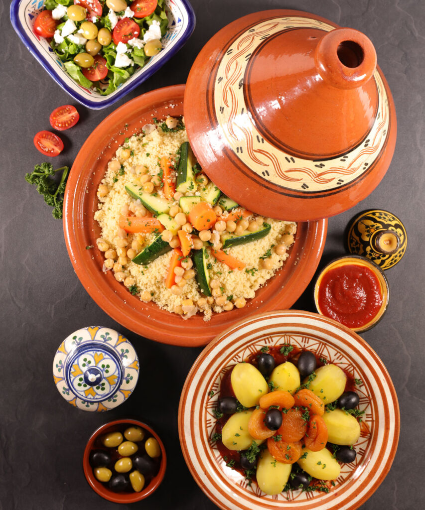 Moroccan meal with couscous and vegetables in a tagine, salad, olives, potatoes with olives, sauce, and decorative bowls on a dark surface.