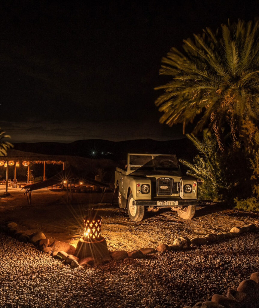 A vintage off-road vehicle is parked at night beside palm trees and lit lanterns, with outdoor seating visible in the background.