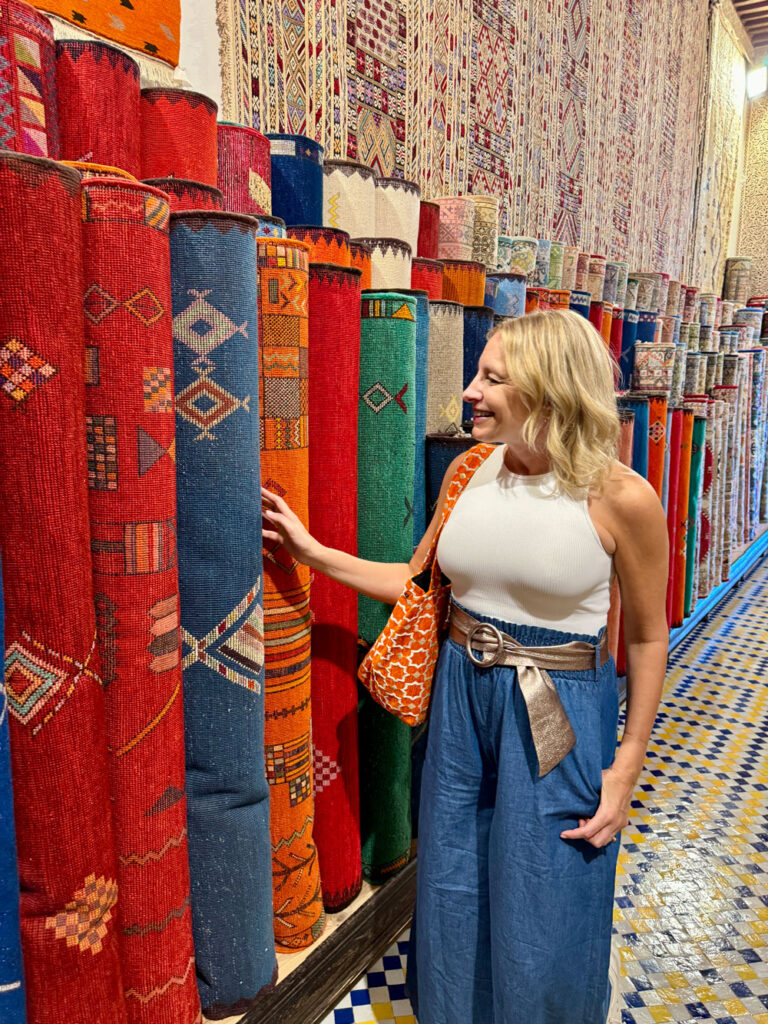 A woman in a white sleeveless top and blue pants stands smiling beside colorful rolled rugs in a shop with patterned floor tiles.
