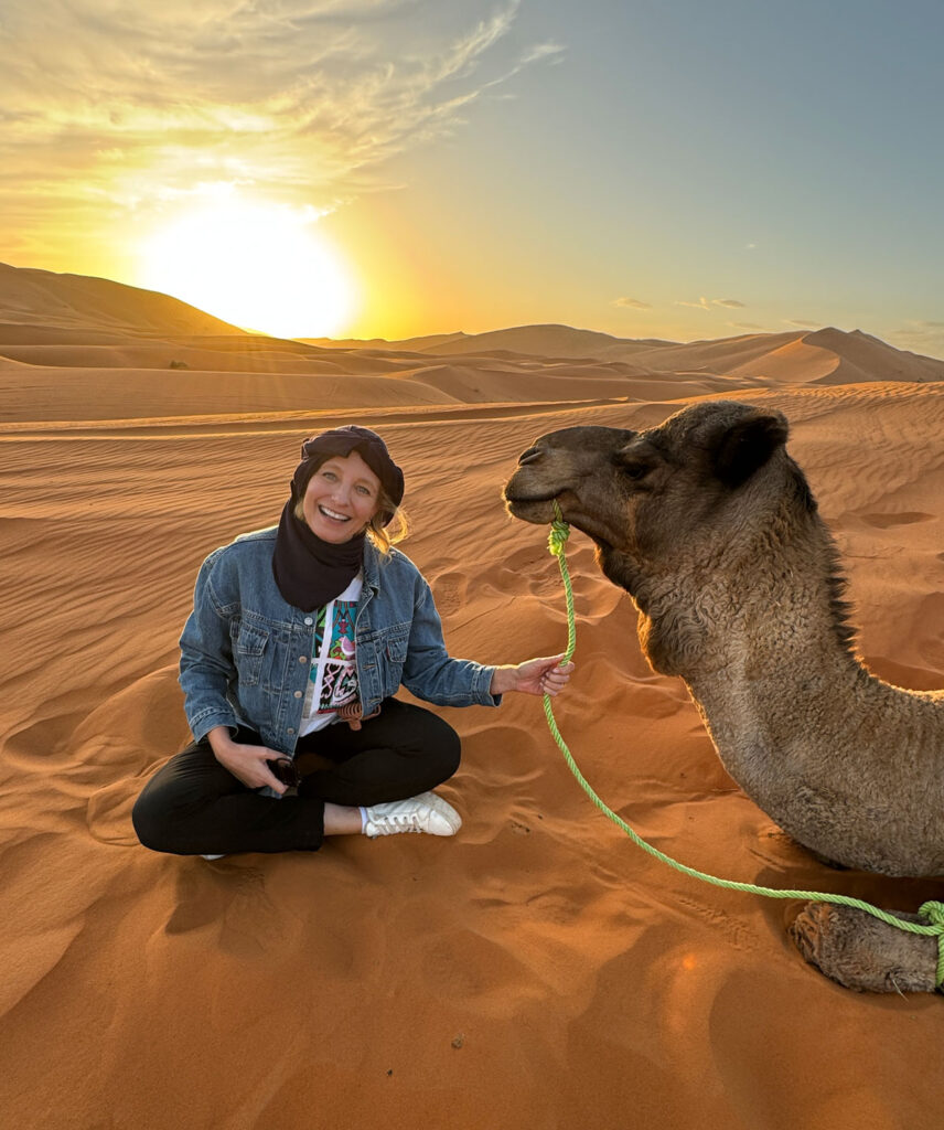 A woman sits on sand next to a camel at sunset in a desert, smiling and holding the camel's lead rope. Rolling sand dunes appear in the background.