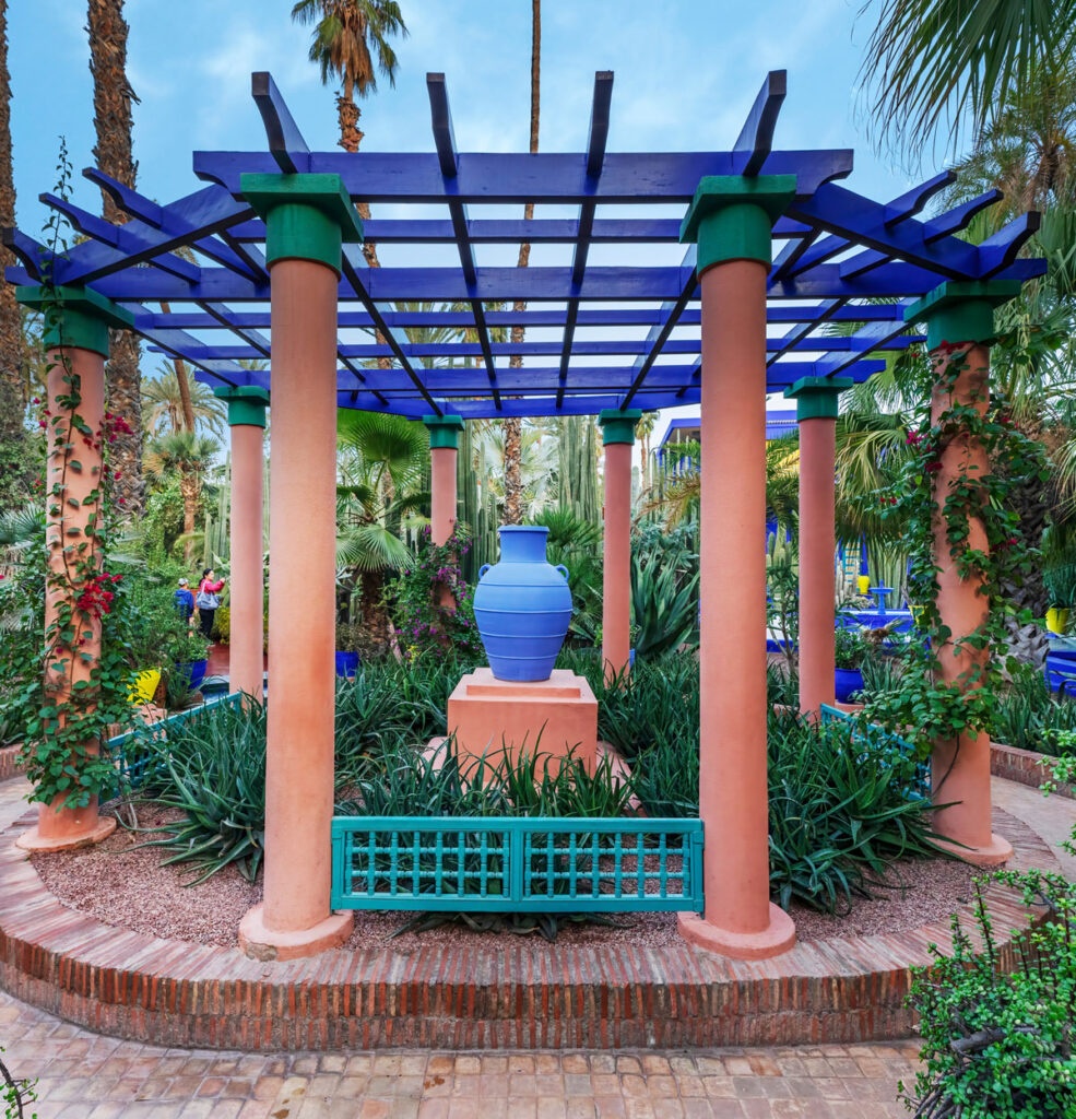 A blue ceramic vase sits on a pedestal under a blue and purple pergola surrounded by lush greenery and tall palm trees in a garden.