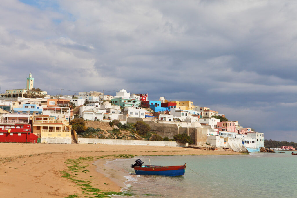 A small wooden boat floats near the shore on a sandy beach, with colorful houses and a mosque visible on a hill under a cloudy sky.
