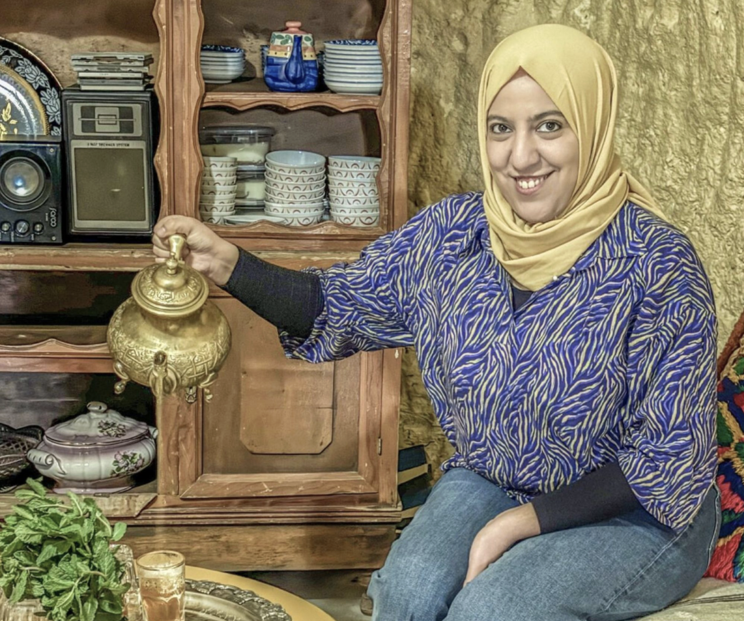 A woman in a hijab smiles while pouring tea from a brass teapot, seated beside a tray with glasses and fresh mint, with dishes and a radio in the background.