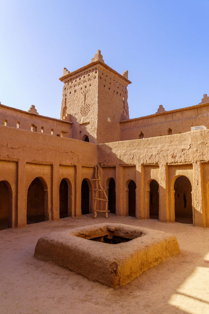 Sunlit courtyard of an adobe building with arches, a central square pit, a wooden ladder, and a tall tower in the background under a clear blue sky.