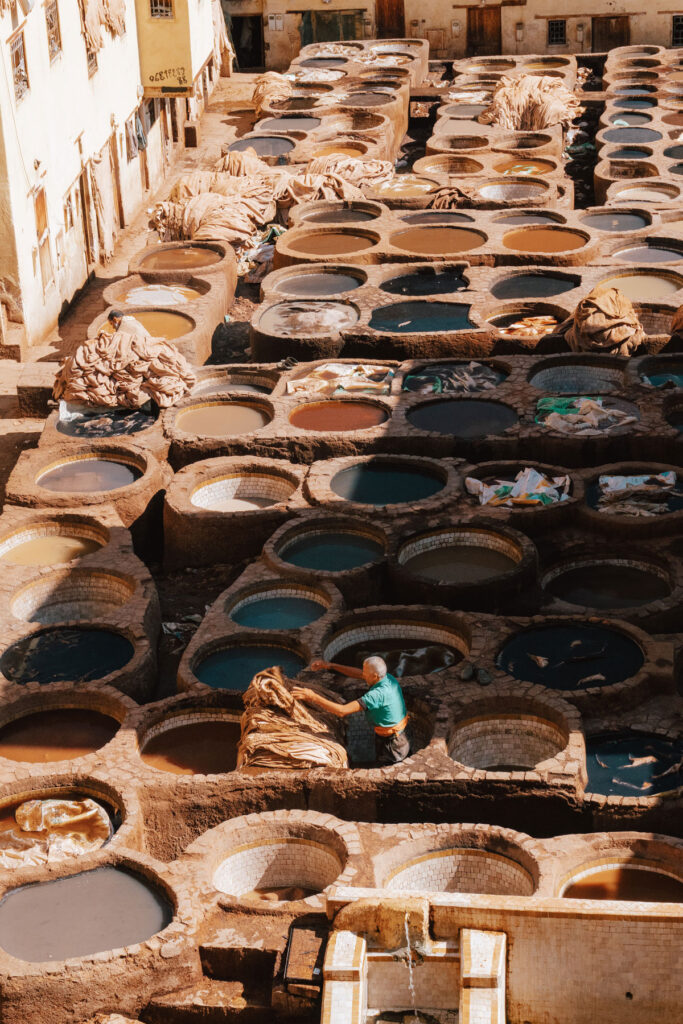 A worker stands among large stone vats filled with various colored liquids at a traditional tannery, surrounded by buildings in sunlight.