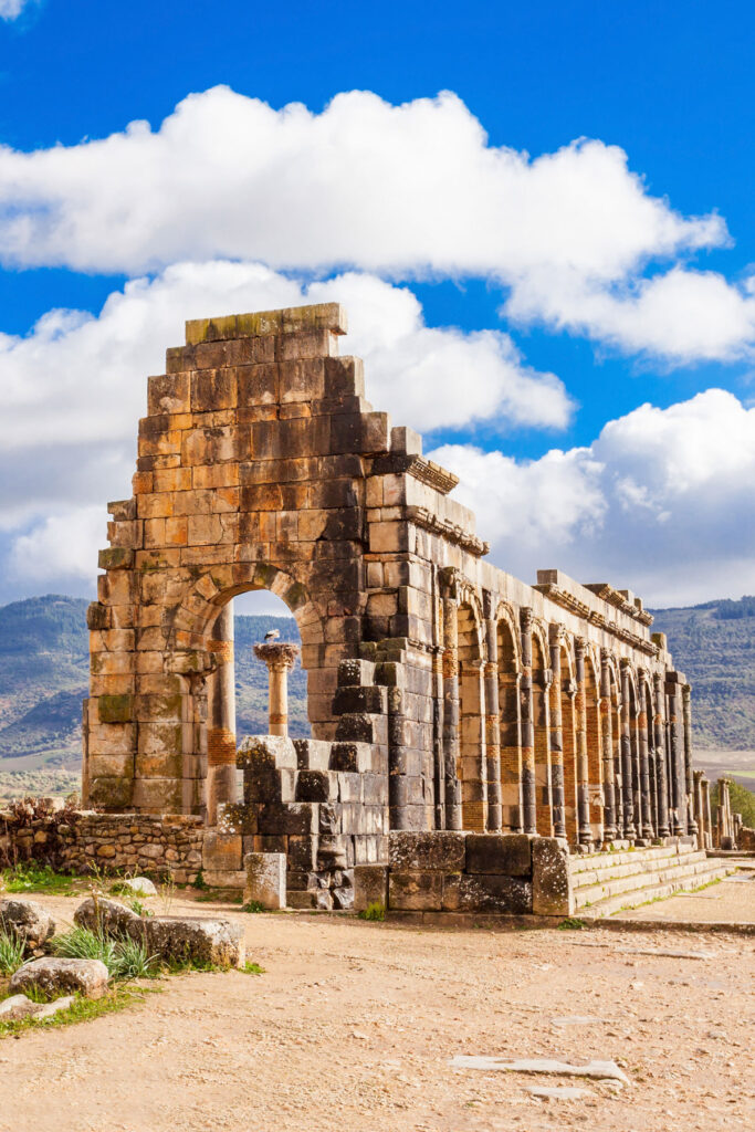 Ancient stone ruins with arches, columns, and partial wall under a blue sky with white clouds, set in an open landscape with hills in the background.