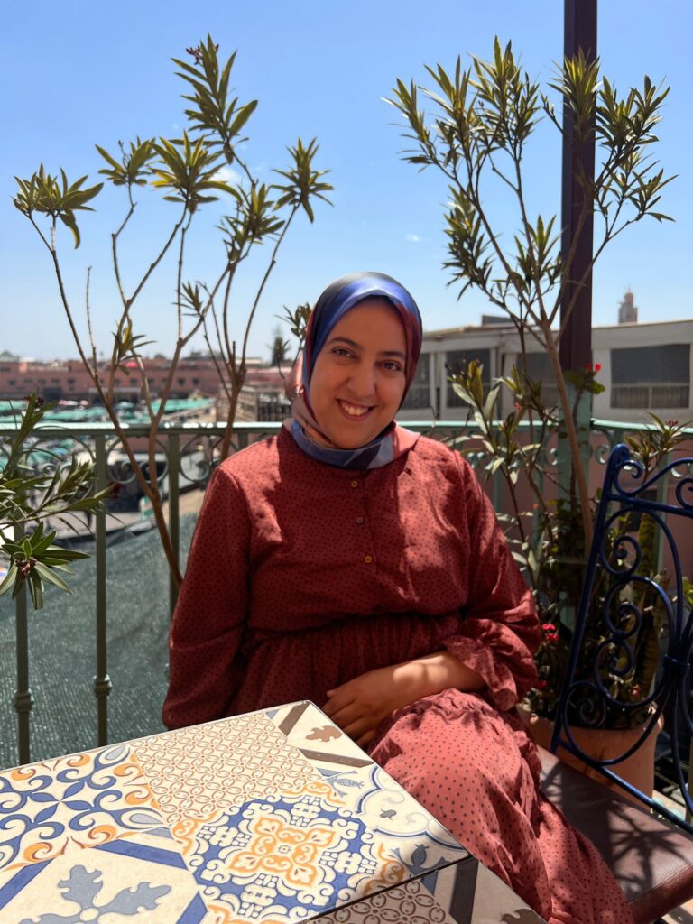 A woman in a red dress and blue headscarf sits at a tiled table on a balcony with plants, under a clear blue sky—perfect inspiration for B2B travel partnerships with Morocco Travel Organizer.
