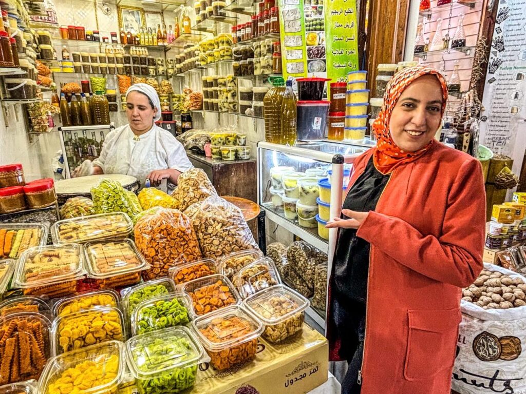 A woman in a red jacket stands in a shop filled with assorted dried fruits, nuts, and sweets, while another woman stands behind the counter.
