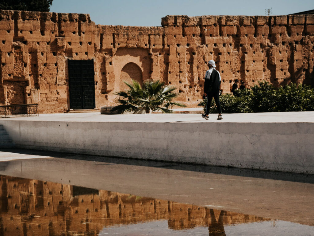 A person walks along a white wall next to a reflective pool, with an ancient, weathered brick structure in the background.