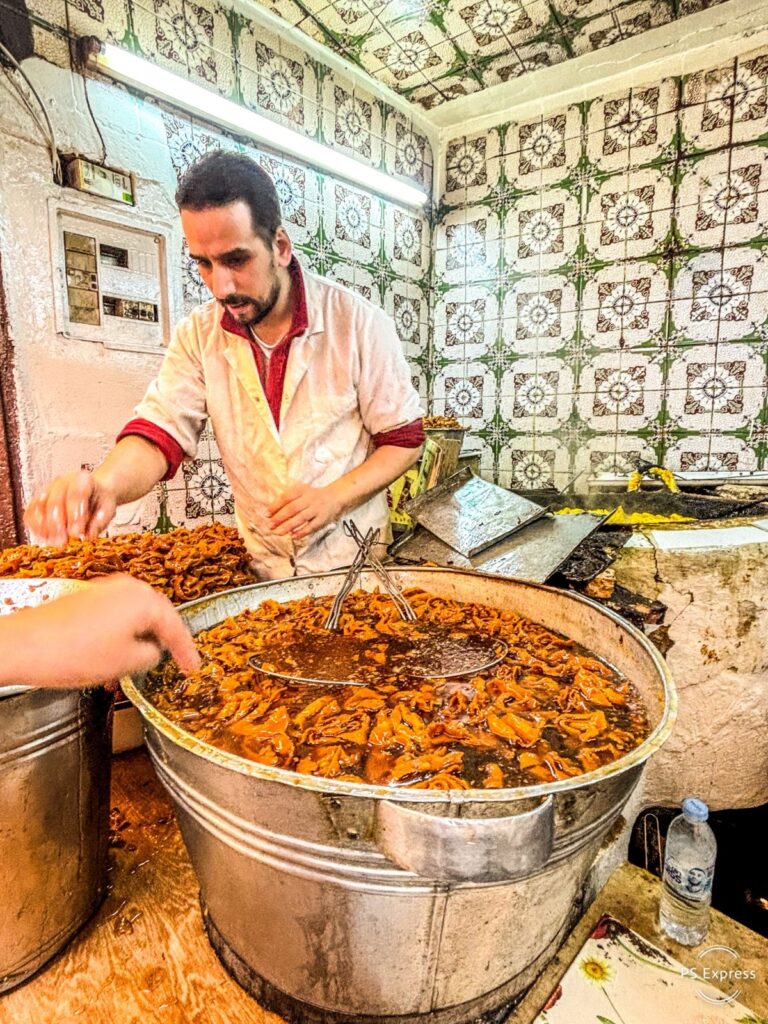 A man in a white coat serves food from a large metal pot filled with a spiced stew in a tiled kitchen.