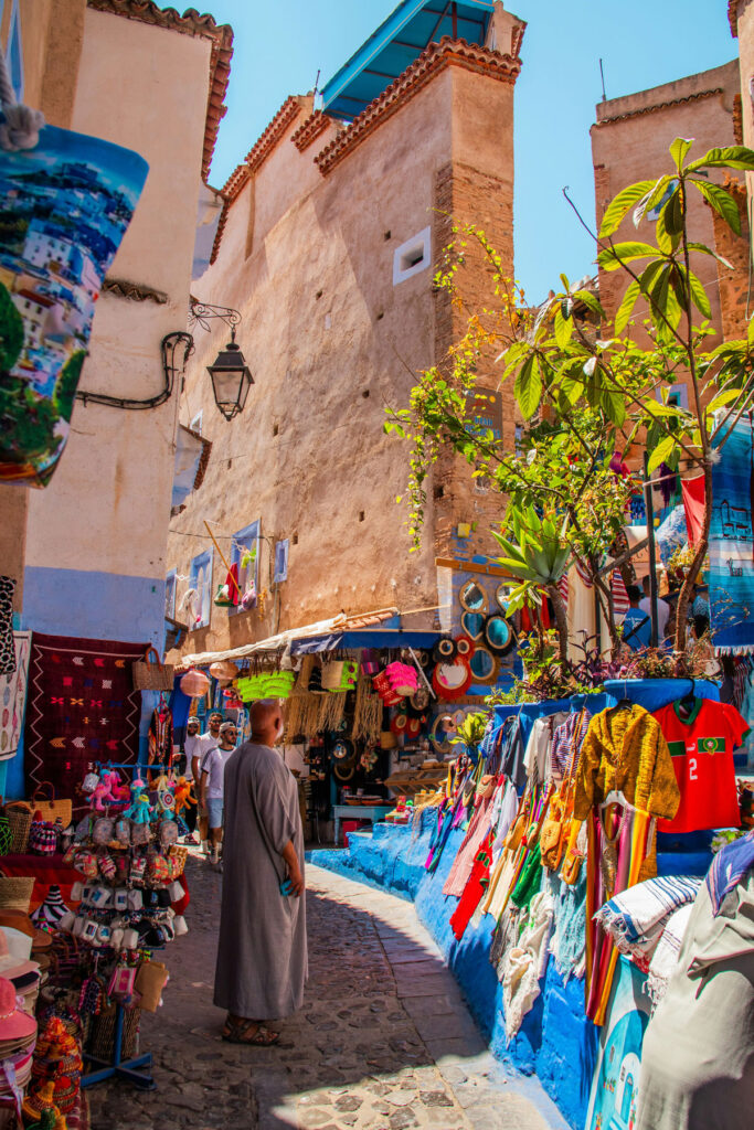 A narrow cobblestone street lined with colorful textiles, clothes, and souvenirs for sale, with a person in a robe walking between tall, sunlit buildings.