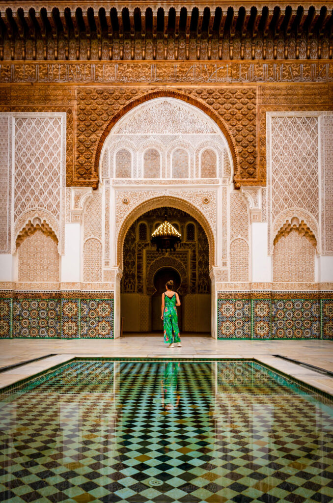 A person in a green dress stands before ornate arches and patterned tiles, reflected in a geometric-tiled pool within a traditional Moroccan-style courtyard—perfect inspiration to plan your luxury culinary or heritage trip to Morocco.