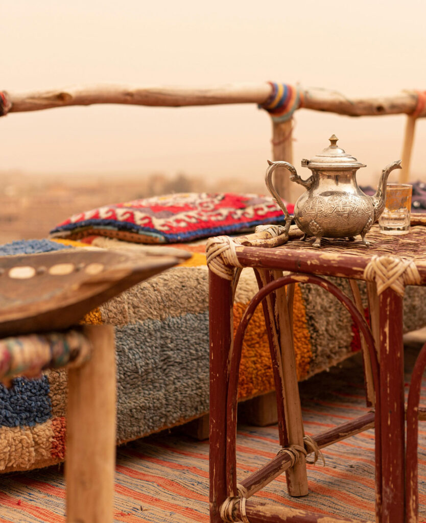 Silver teapot and glass on a wooden table outdoors, with colorful woven cushions and blankets on benches, set against a hazy, desert-like background.