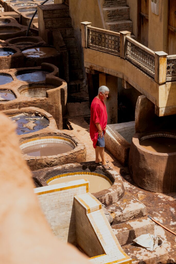 A person in a red shirt walks among circular stone vats at a traditional tannery under direct sunlight.