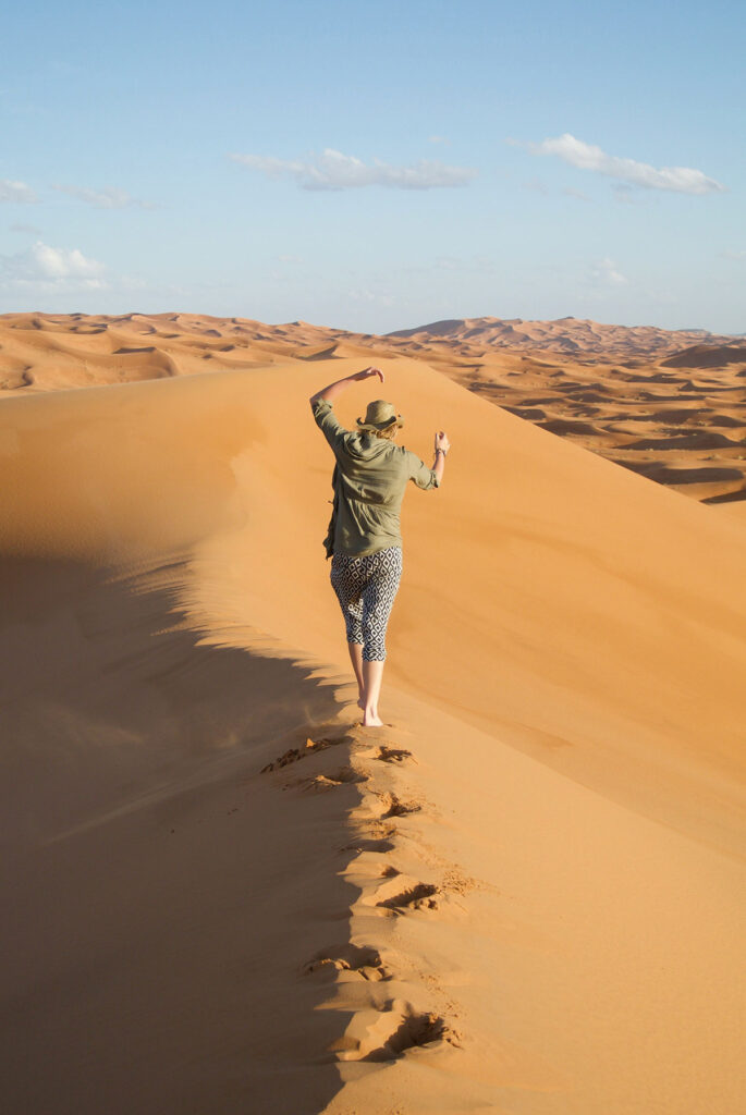 Person walks barefoot along a sand dune ridge in a vast desert landscape under a clear sky, leaving footprints behind.